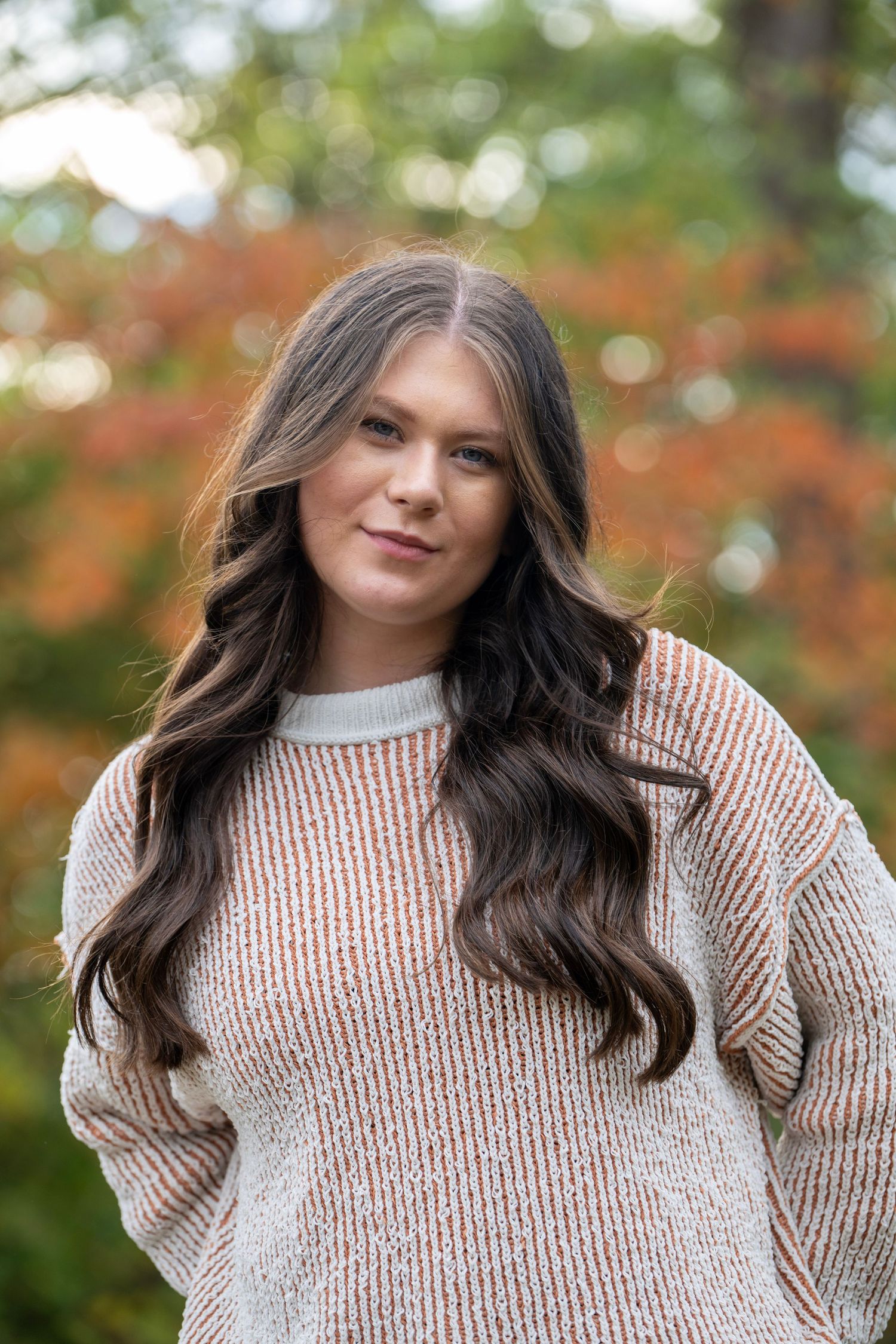 Person in cozy beige sweater poses outdoors with fall foliage in background.