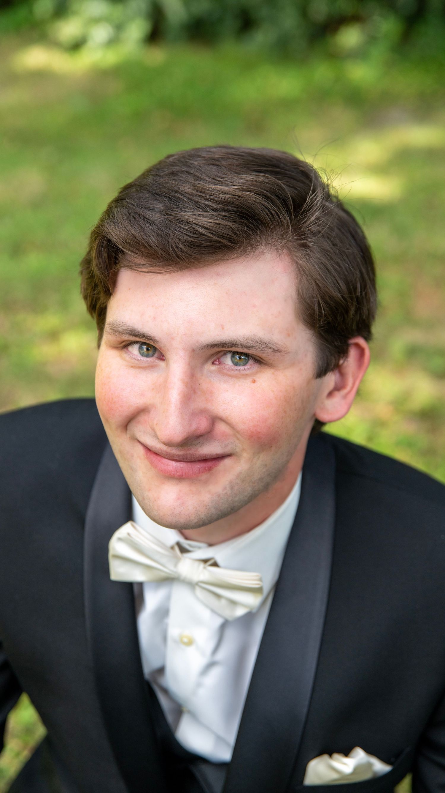 Person in formal black tuxedo and white bow tie smiling in an outdoor garden setting.