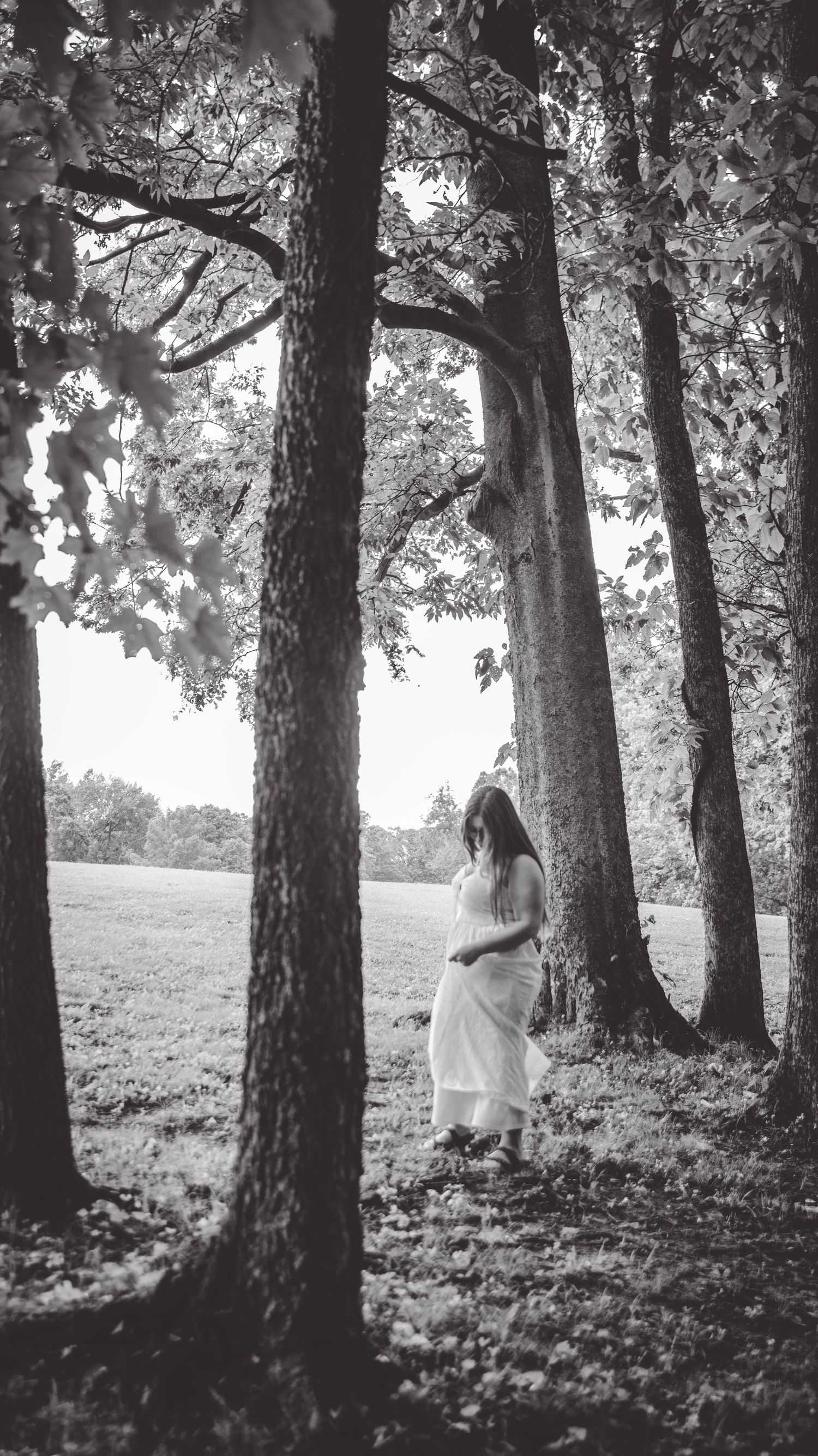 Black and white portrait of a person in white dress among forest trees.