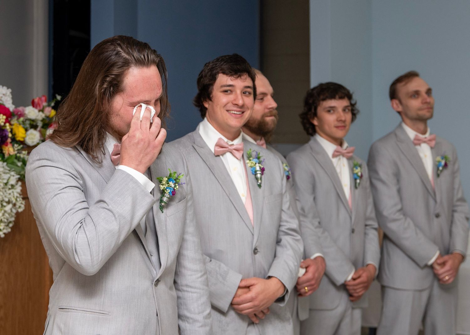 Wedding party members in light gray suits show emotional reactions during the ceremony.