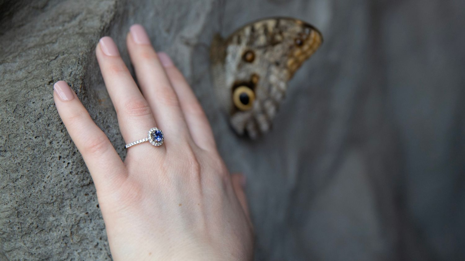 A delicate engagement ring with a blue stone photographed with a butterfly in the background.