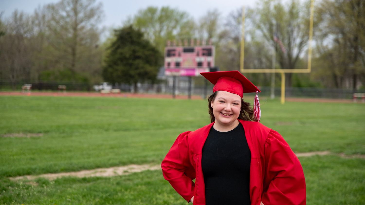 Graduate in red regalia poses on a running track surrounded by spring greenery.