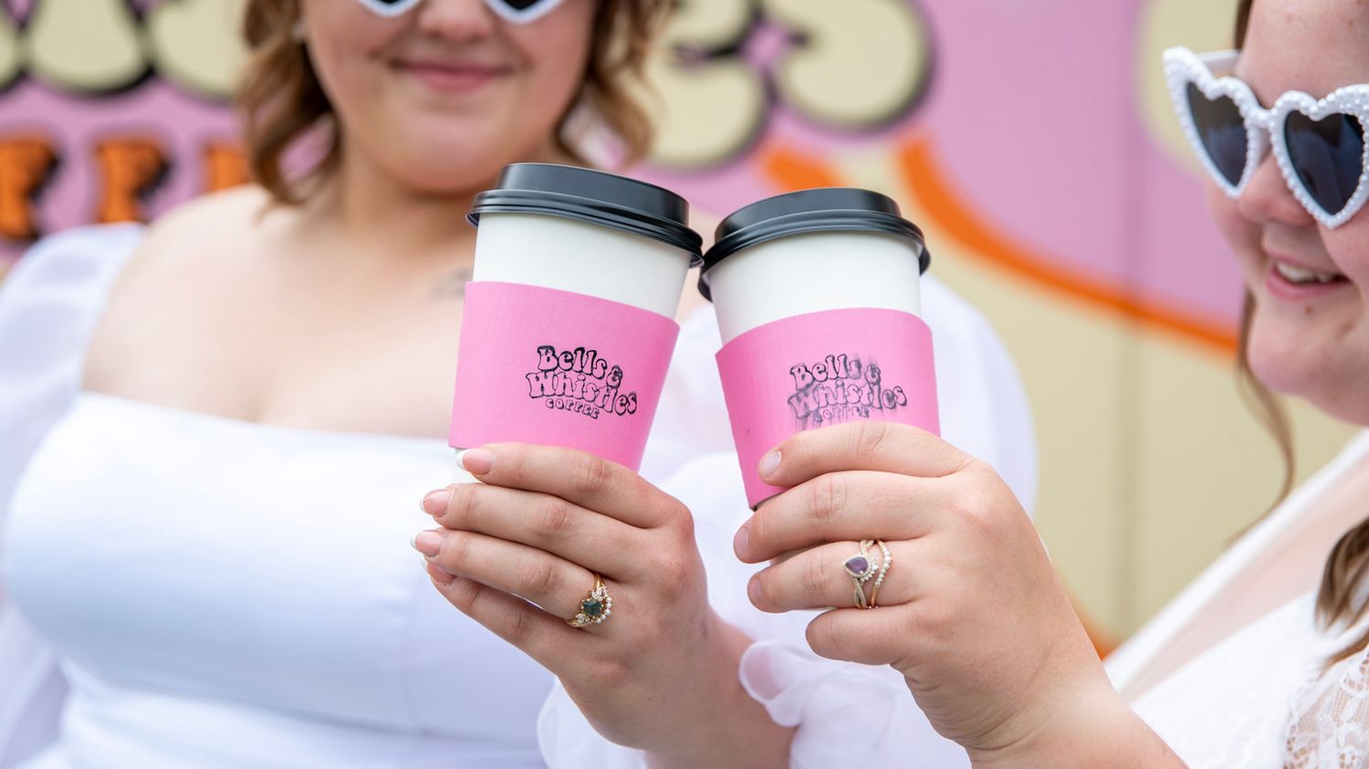 Pink coffee cups held by hands wearing rings against blurred storefront background.
