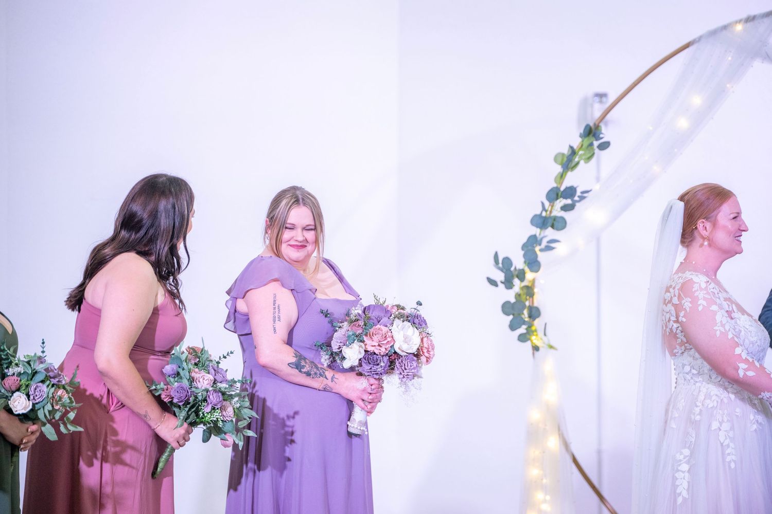 Bridesmaids in purple and mauve dresses hold bouquets beside a decorative circular arch at a wedding ceremony.