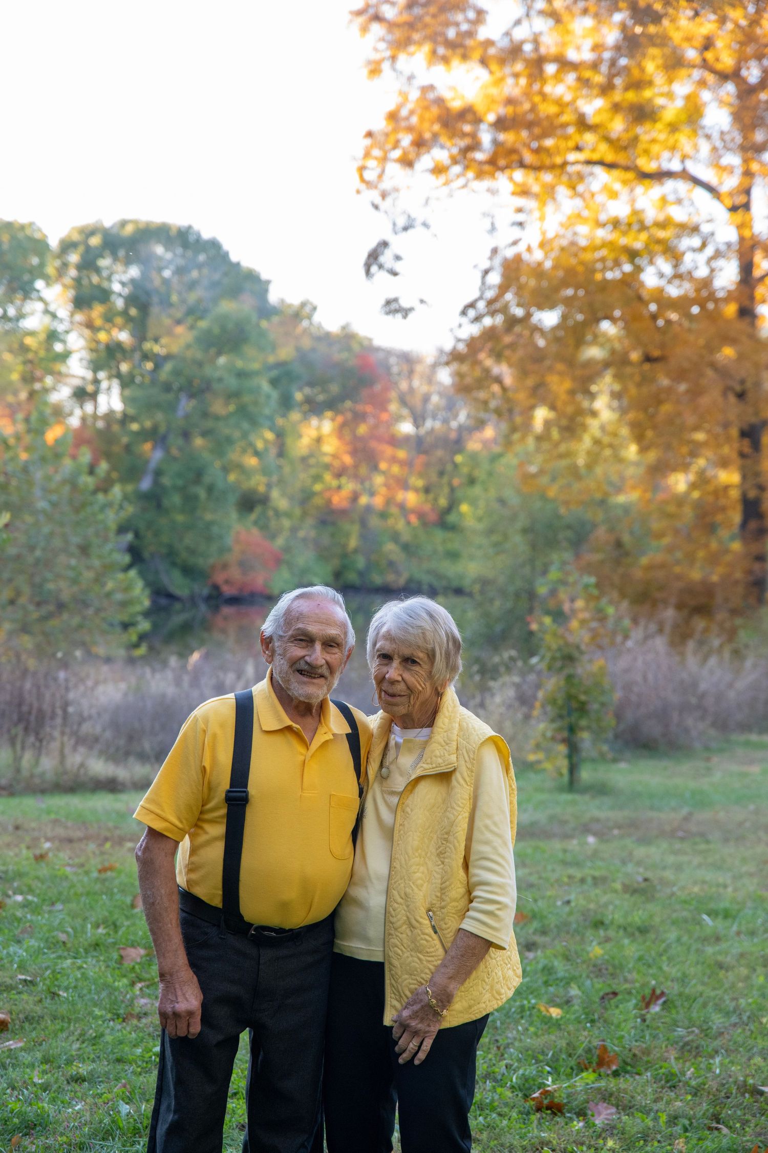 Senior couple in matching yellow attire pose together in a fall landscape with golden trees.