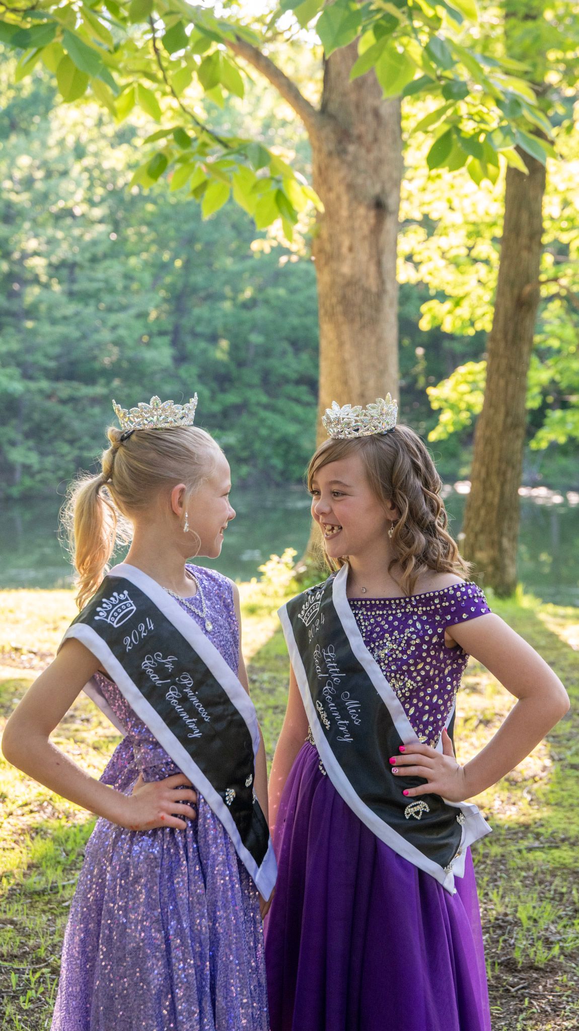 Beauty queens in sparkling purple dresses pose together under sunlit trees.