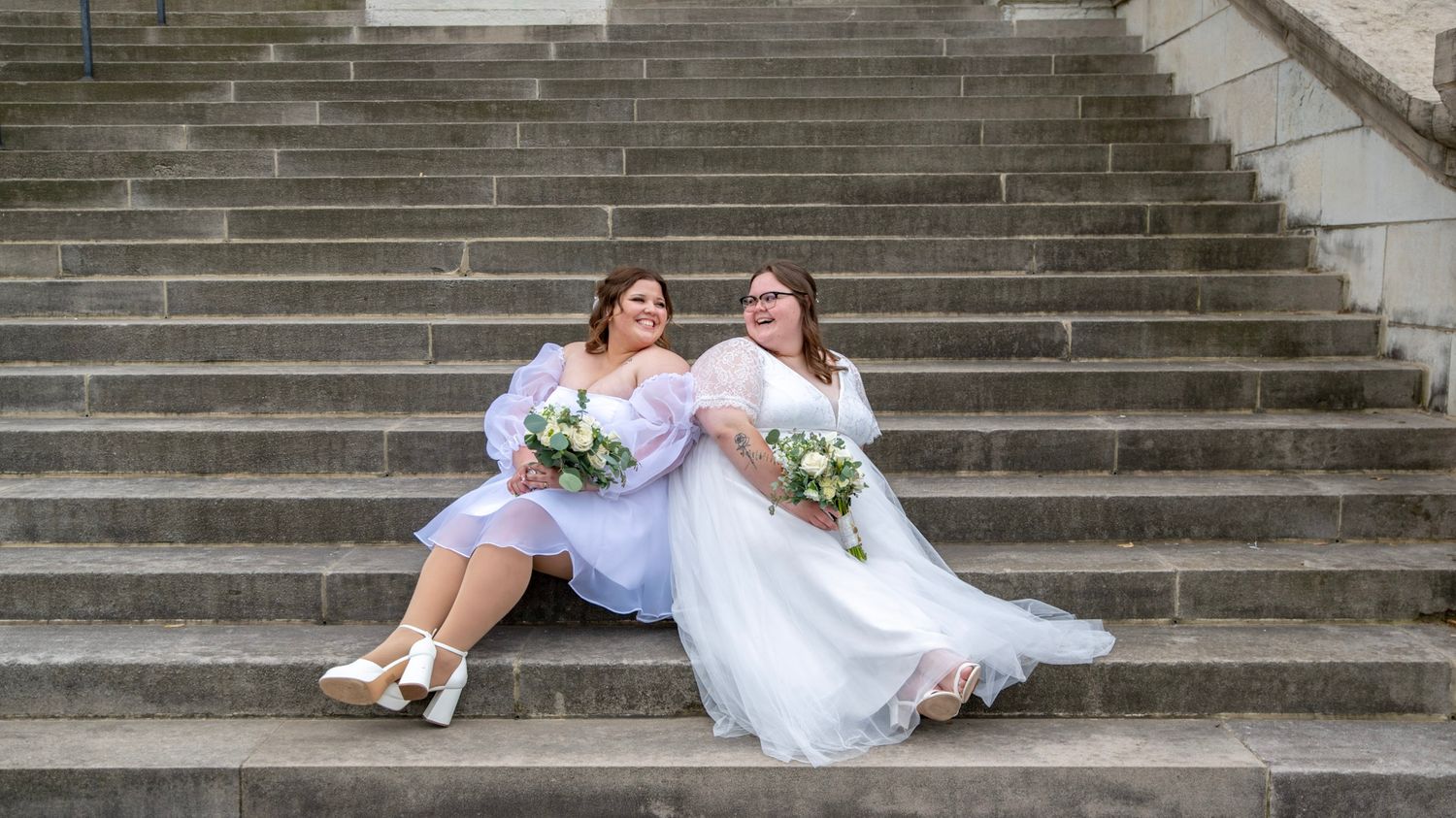 Couple in wedding attire pose playfully on wide stone staircase.