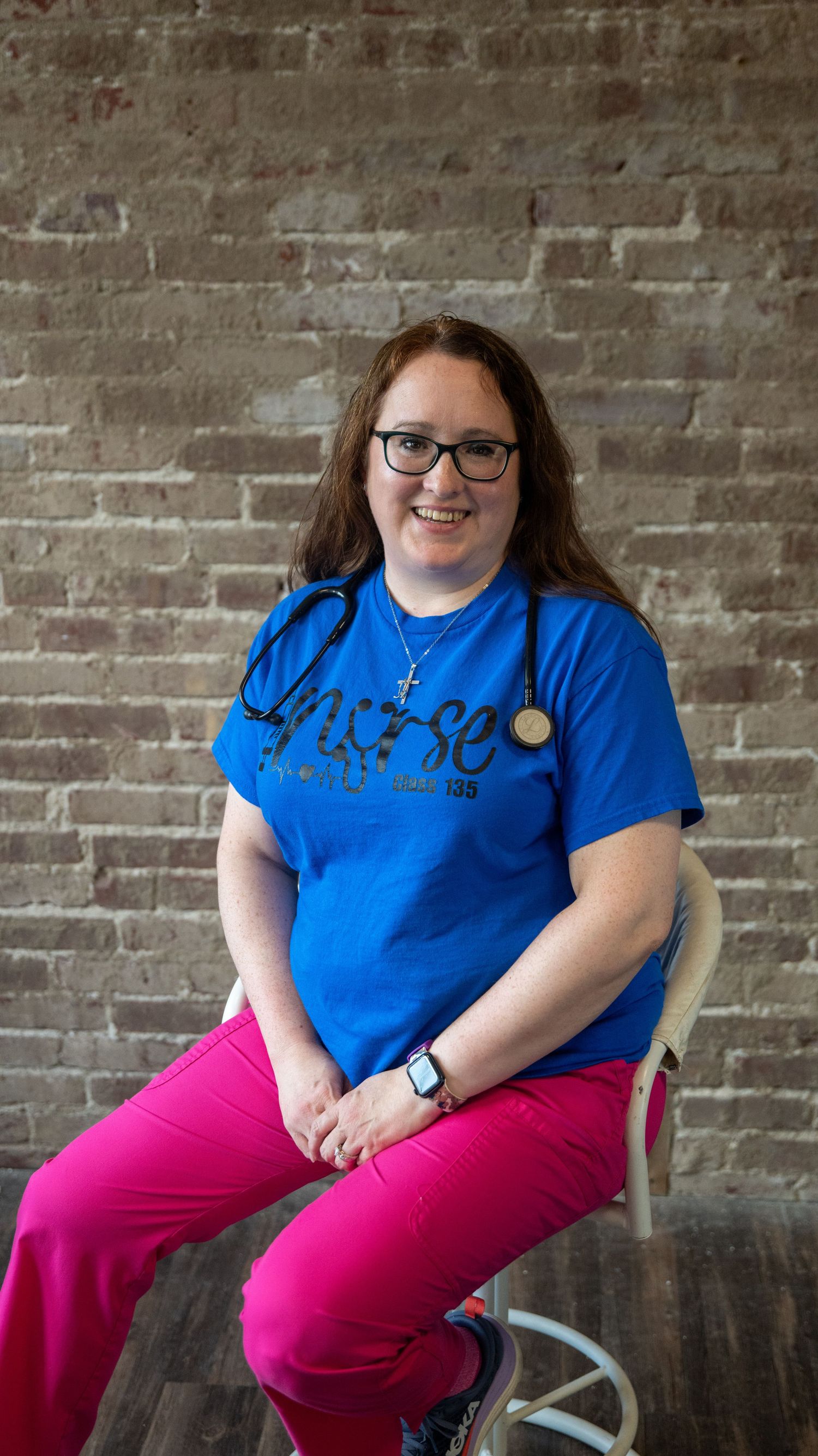 A person in glasses wearing blue and pink clothing sits casually against a rustic brick wall background.