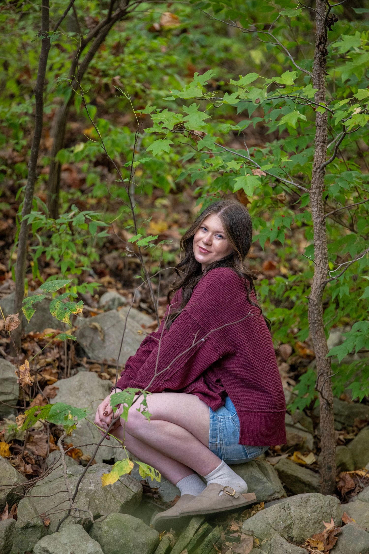 Person in burgundy sweater sits on rocky terrain surrounded by green foliage.