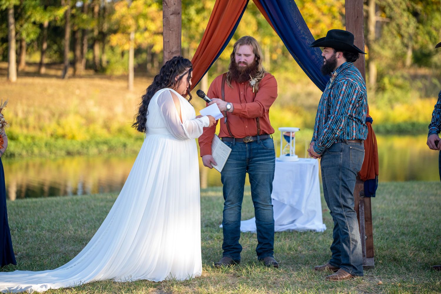 Wedding party poses outdoors with rustic wooden arch decorated in navy and rust-colored draping at sunset.