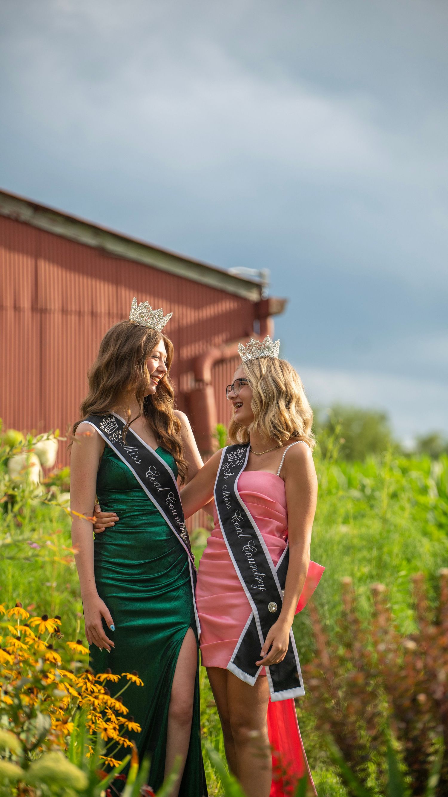 Crowned winners in green and pink formal dresses share a moment in garden setting.