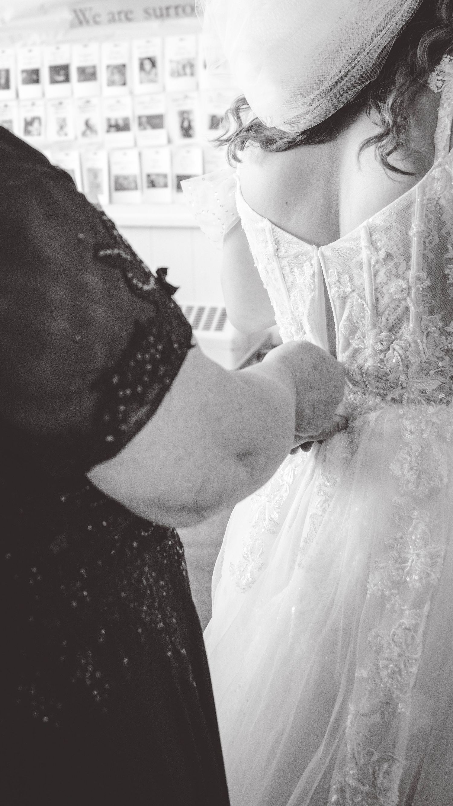 Close-up black and white photo showing a wedding dress being adjusted in front of a wall with photographs.
