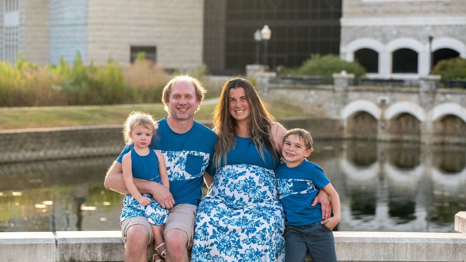 Family photoshoot by a scenic stone bridge and pond wearing matching blue outfits.