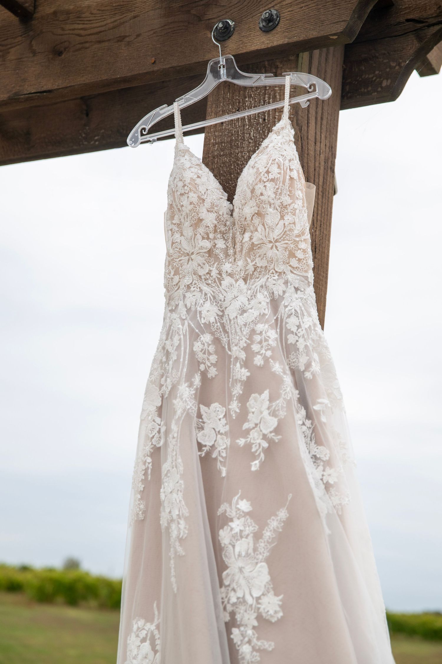 Elegant white lace wedding dress with floral embroidery details hanging on display against a rustic wooden backdrop.