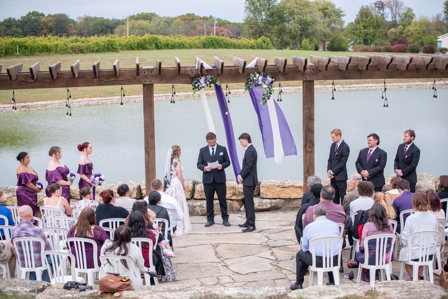 Lakeside wedding ceremony with purple decor and white chairs under wooden pergola.