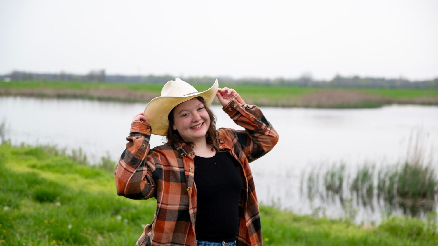 Person in plaid shirt and cowboy hat poses near wetlands.
