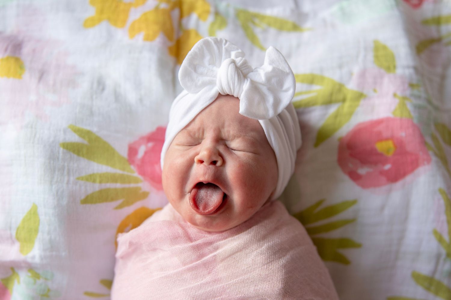 Baby wearing white bonnet makes adorable expression on floral patterned fabric.