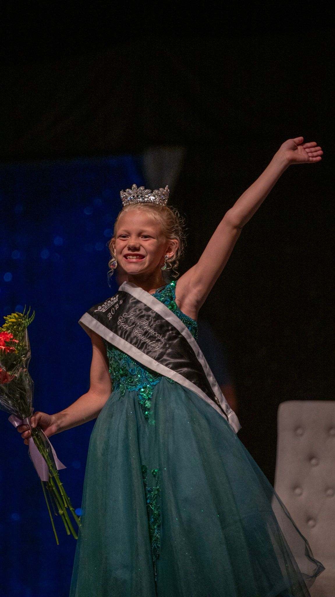 A pageant contestant in a sparkly teal gown and tiara celebrates on stage with raised arms.
