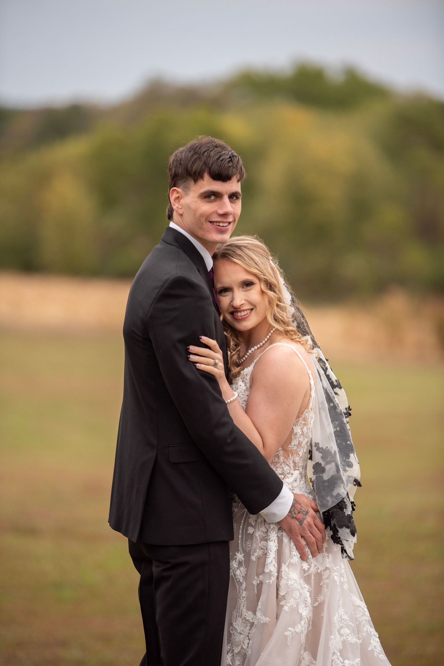 A romantic wedding moment captured in a sunlit field with long white dress and black suit.