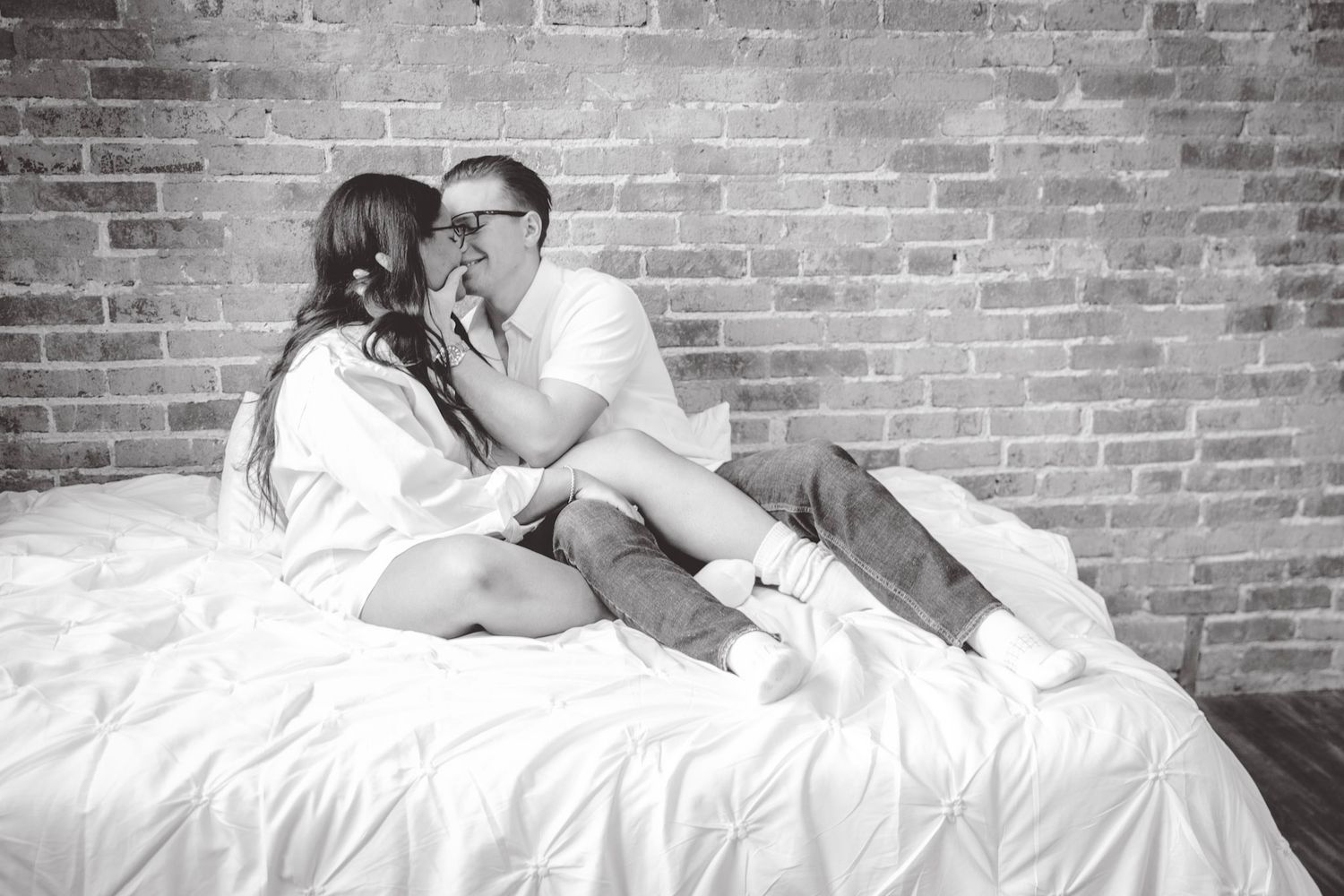 Couple sharing tender moments on white bed with brick wall backdrop in monochrome.