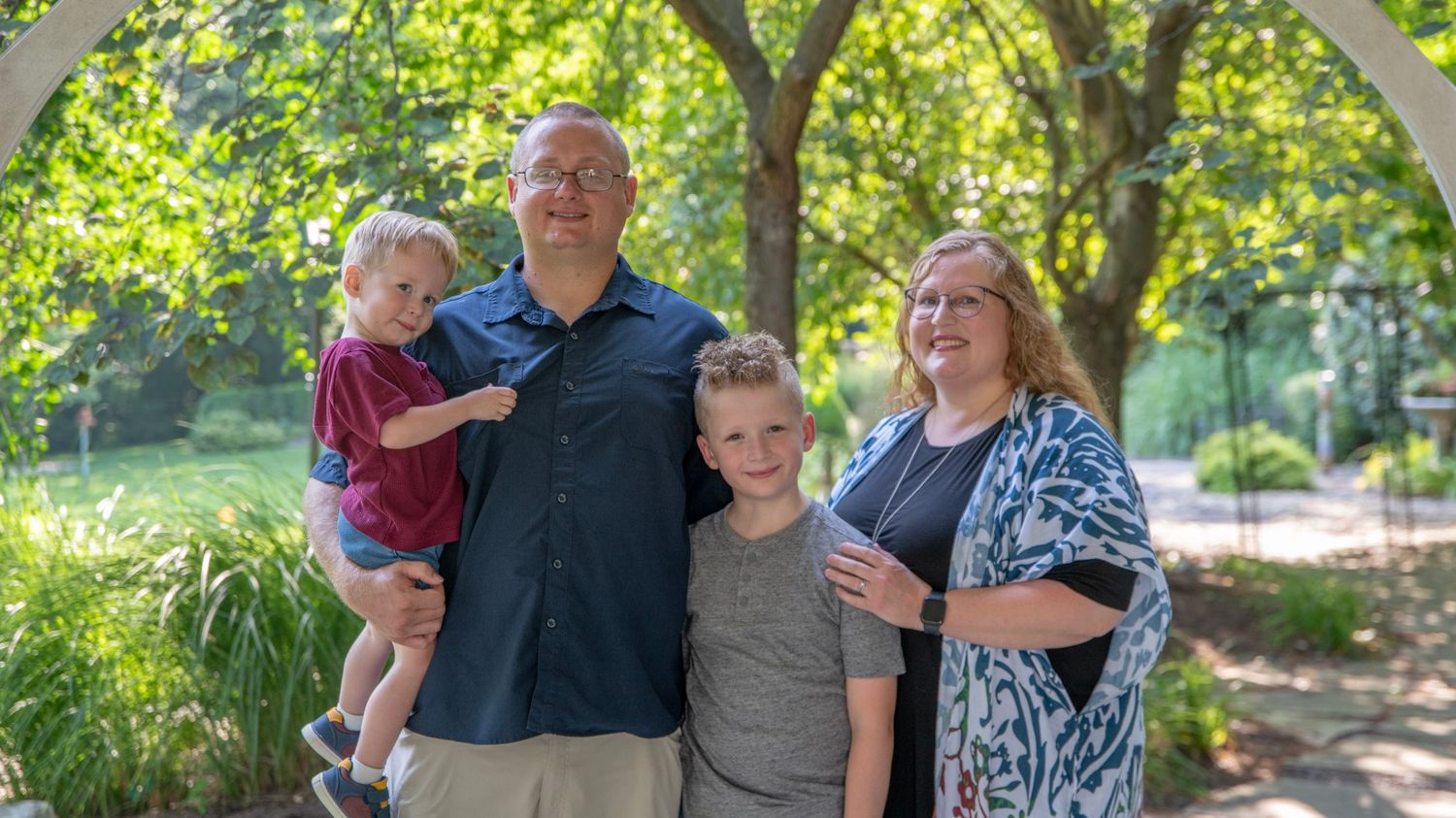 Family members sitting together on a park bench surrounded by lush greenery on a sunny day.