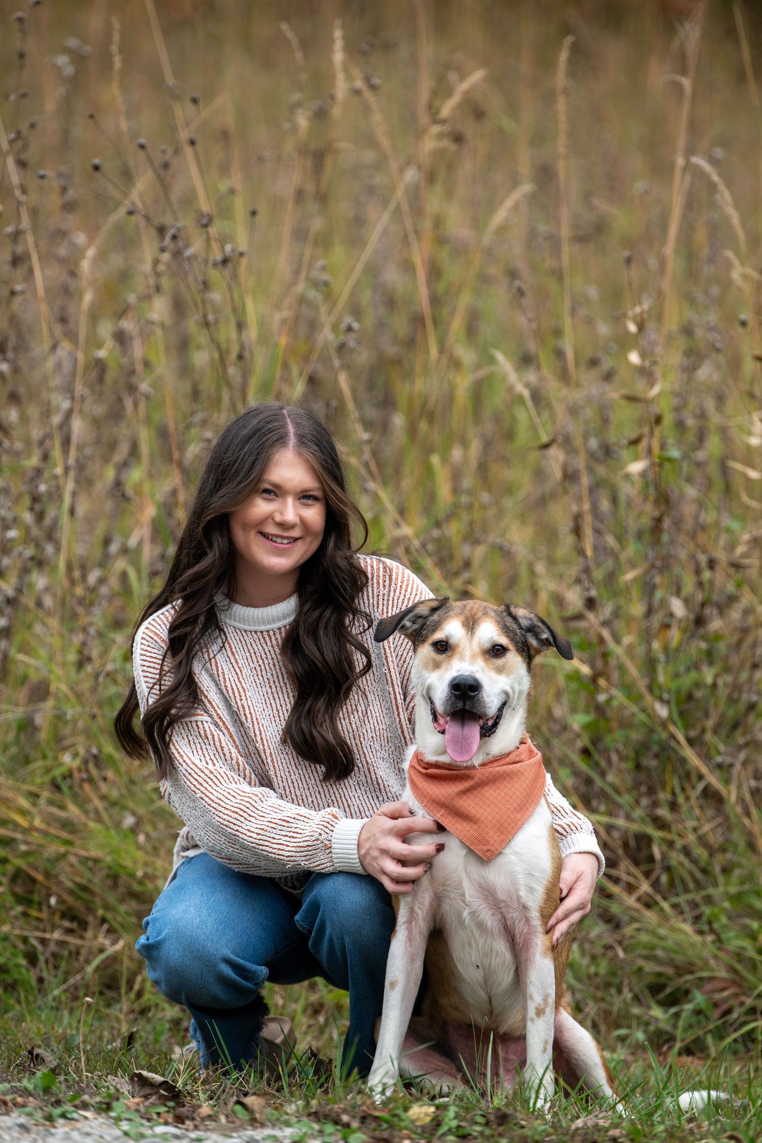 A mixed breed dog wearing a bandana poses with their owner in a field of tall grass.