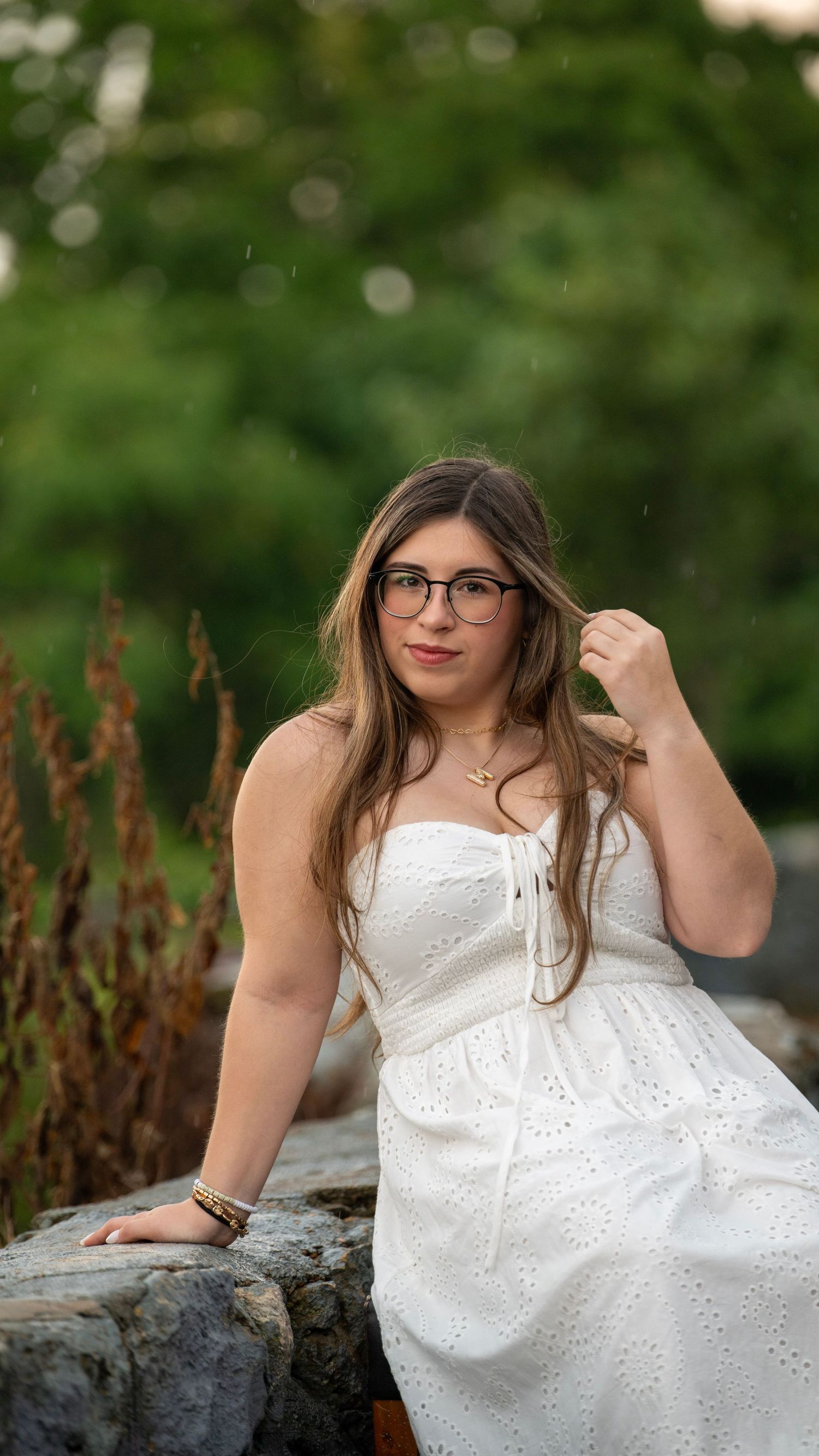 Model in white eyelet dress reclines on stone wall with green background.