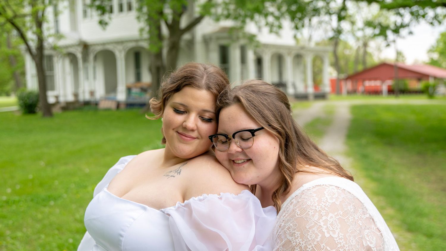 Two people in wedding dresses embrace on a lawn with historic buildings in the background.