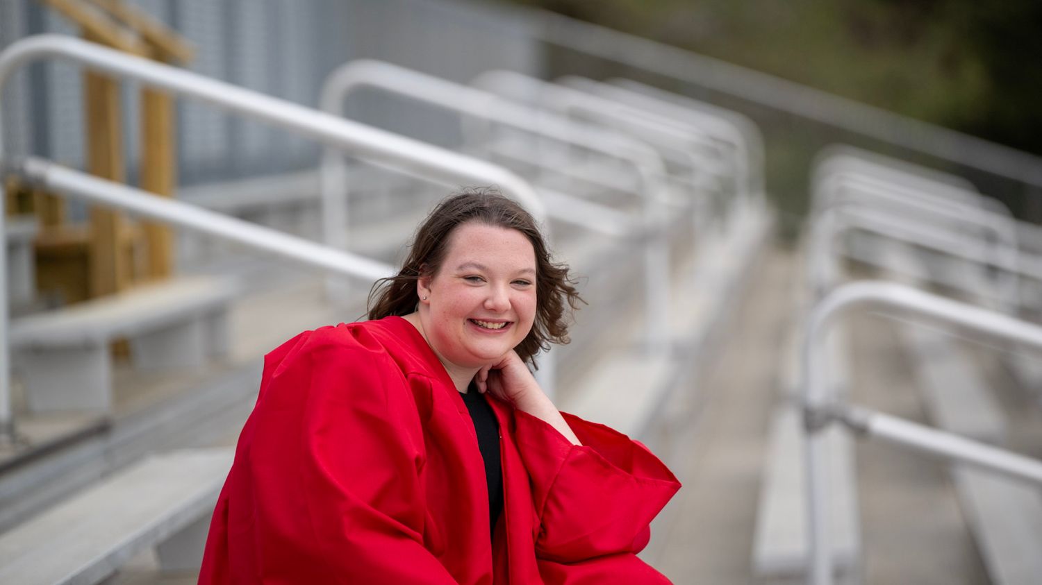 Graduate in red cap and gown poses on bleachers with metal railings.