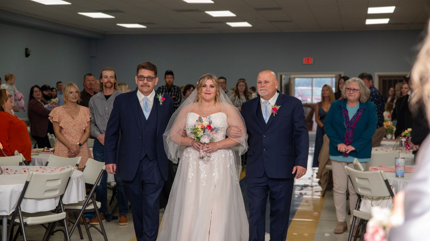 A wedding processional in a reception hall with white chairs and guests looking on.