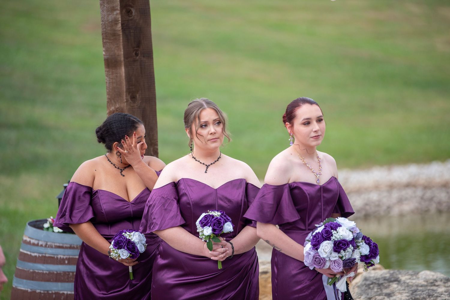 Three bridesmaids in purple off-shoulder dresses hold bouquets while standing outdoors.