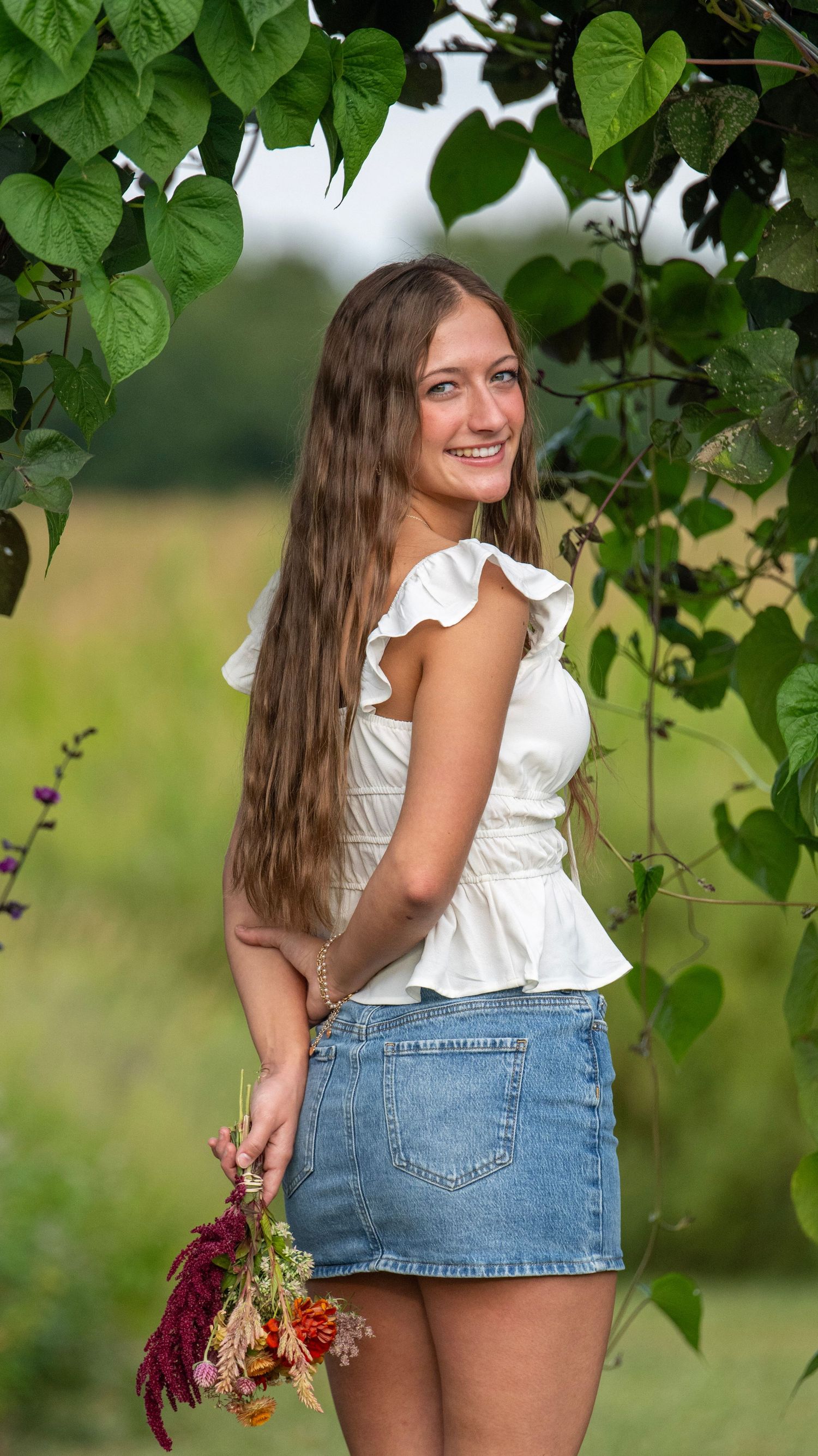 A person in white ruffled top and denim skirt holds dried flowers under a garden arch.