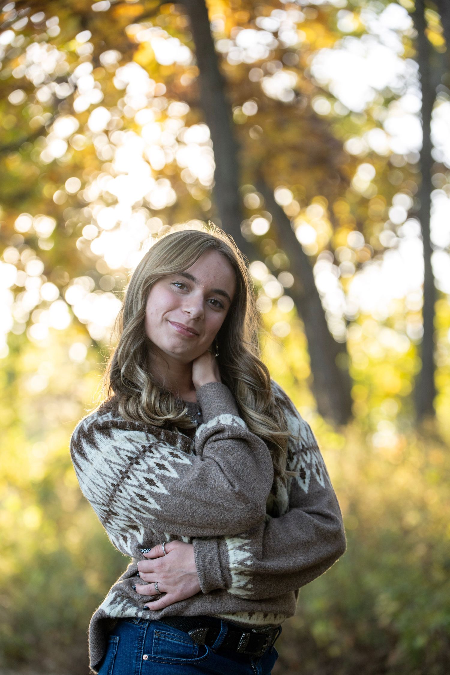 A person in a gray sweater and jeans poses outdoors with autumn trees and sunlight creating a warm atmosphere.