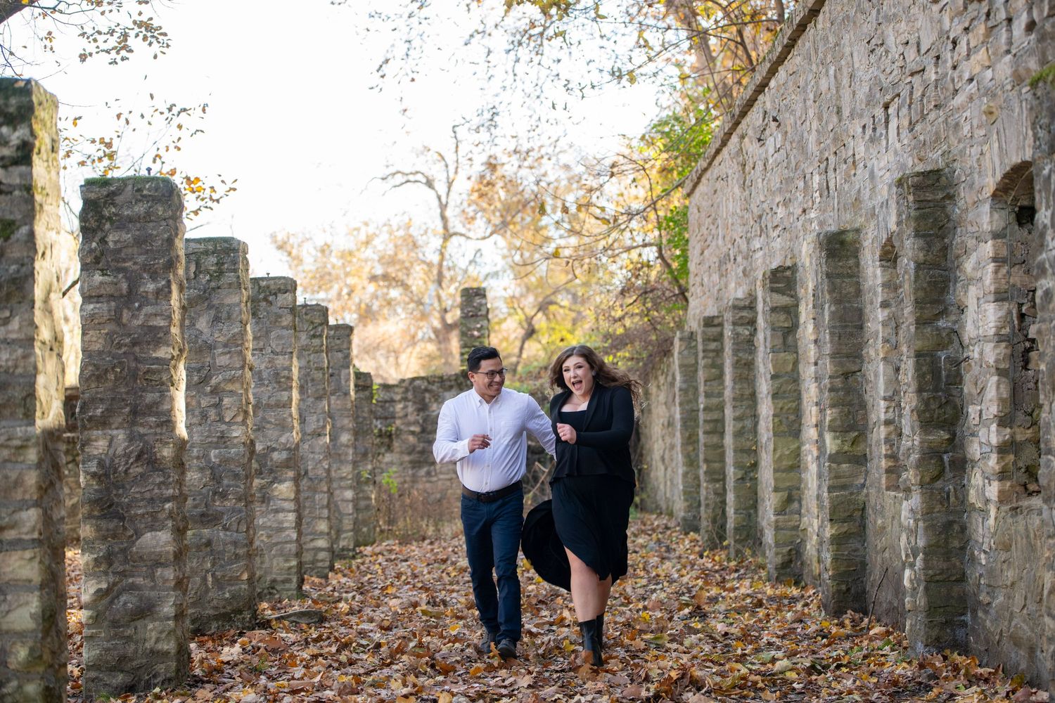 A romantic couple poses against stone pillars in an autumn setting with fallen leaves.