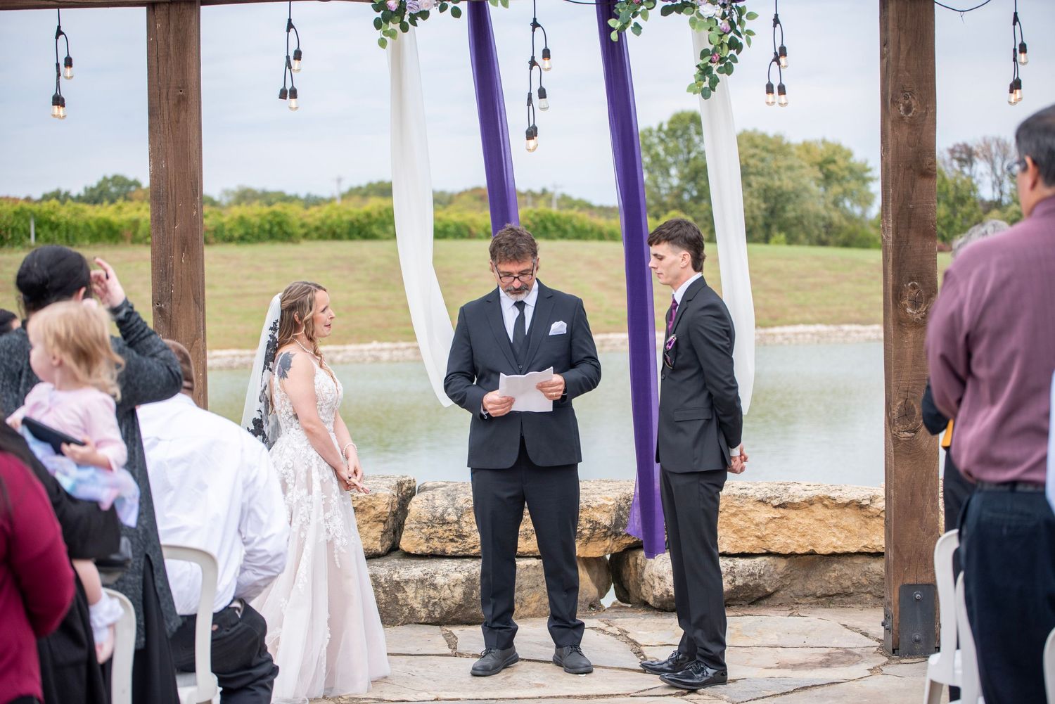 Wedding ceremony under wooden pergola with lake and greenery in background.