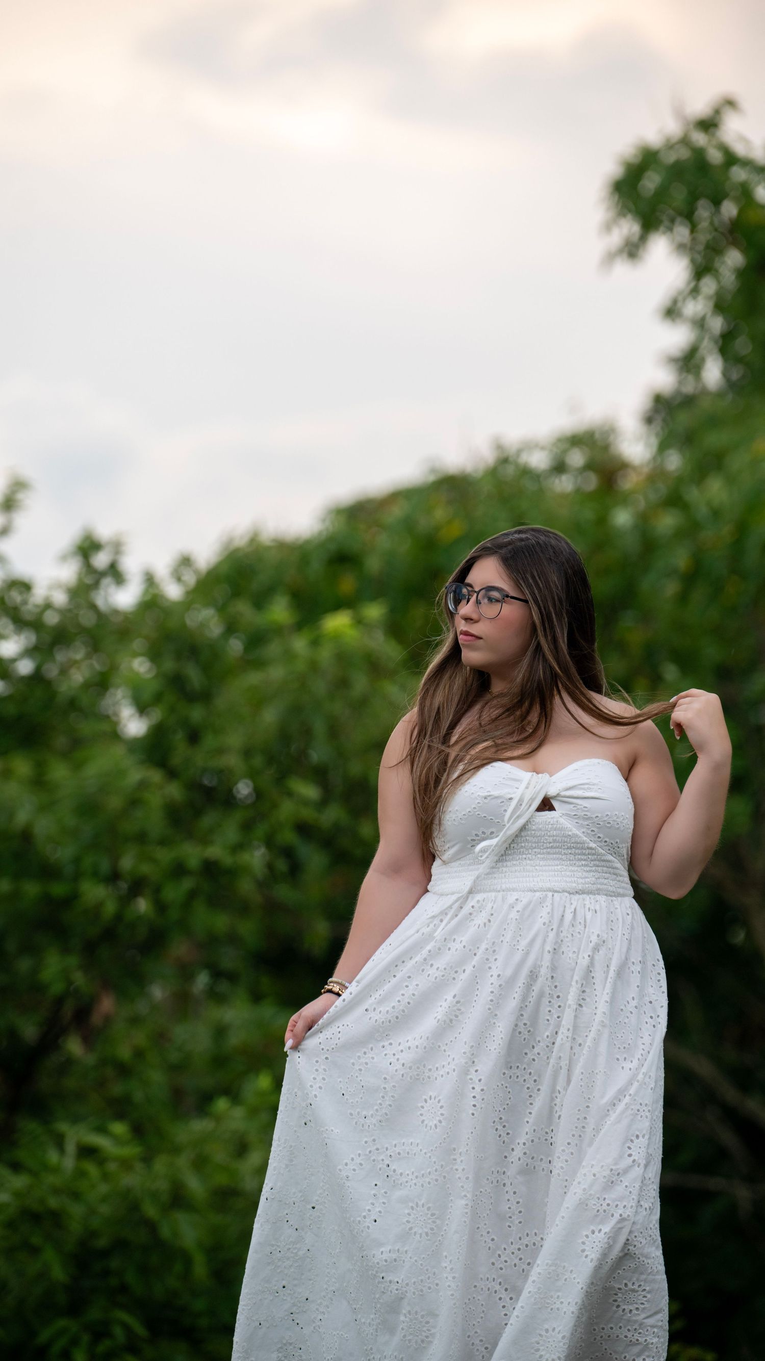 A person in glasses and white dress poses outdoors against green foliage.