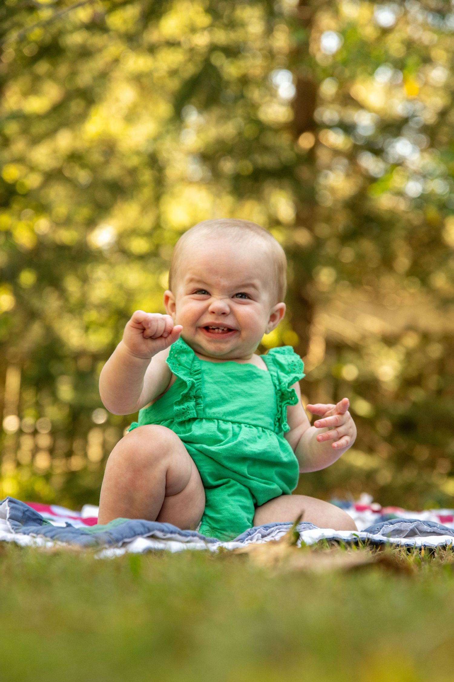 Joyful baby in green romper sitting and laughing on blanket in dappled sunlight.