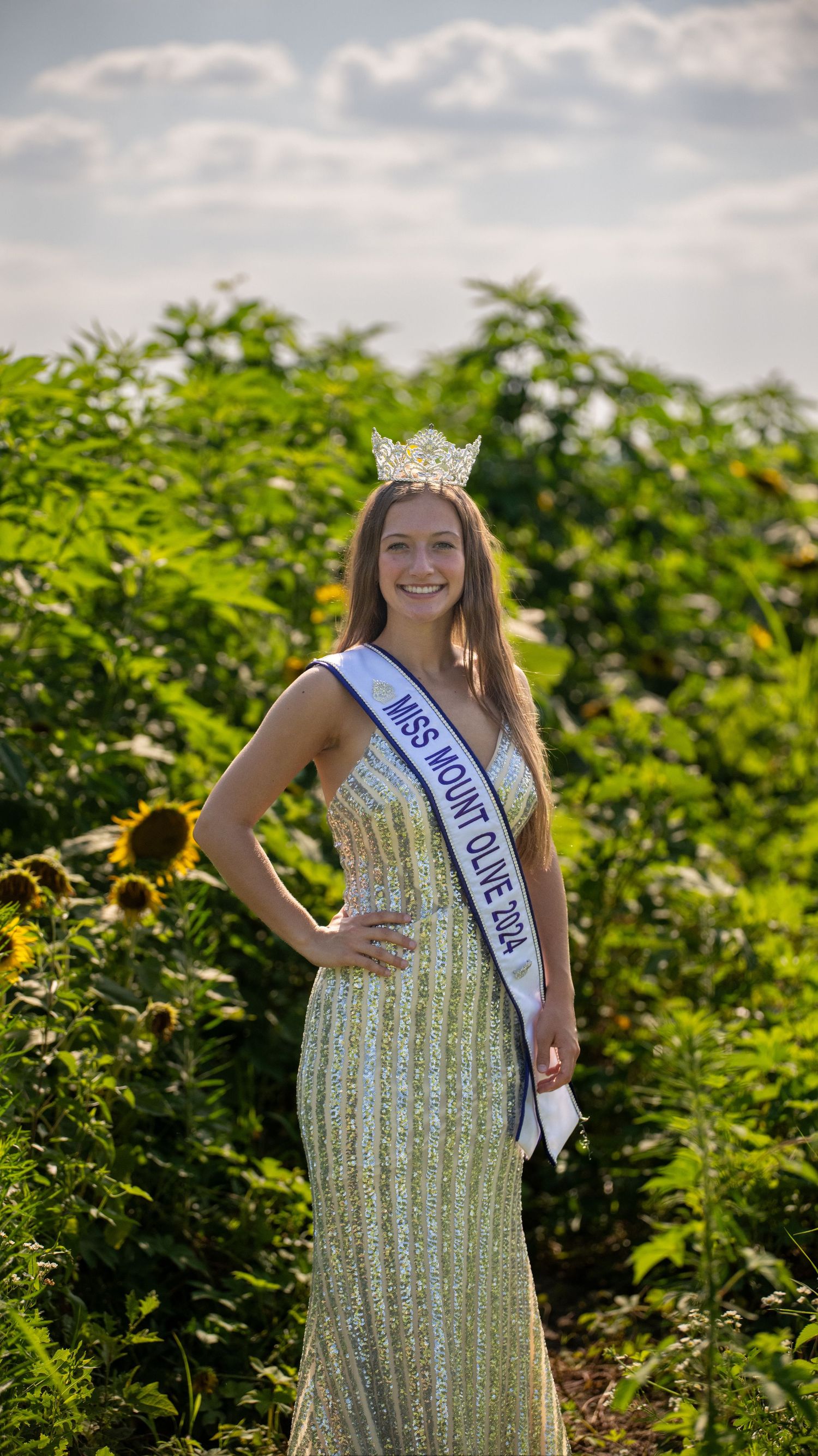 A pageant winner in a sparkly dress poses in a sunflower field wearing a crown and sash.