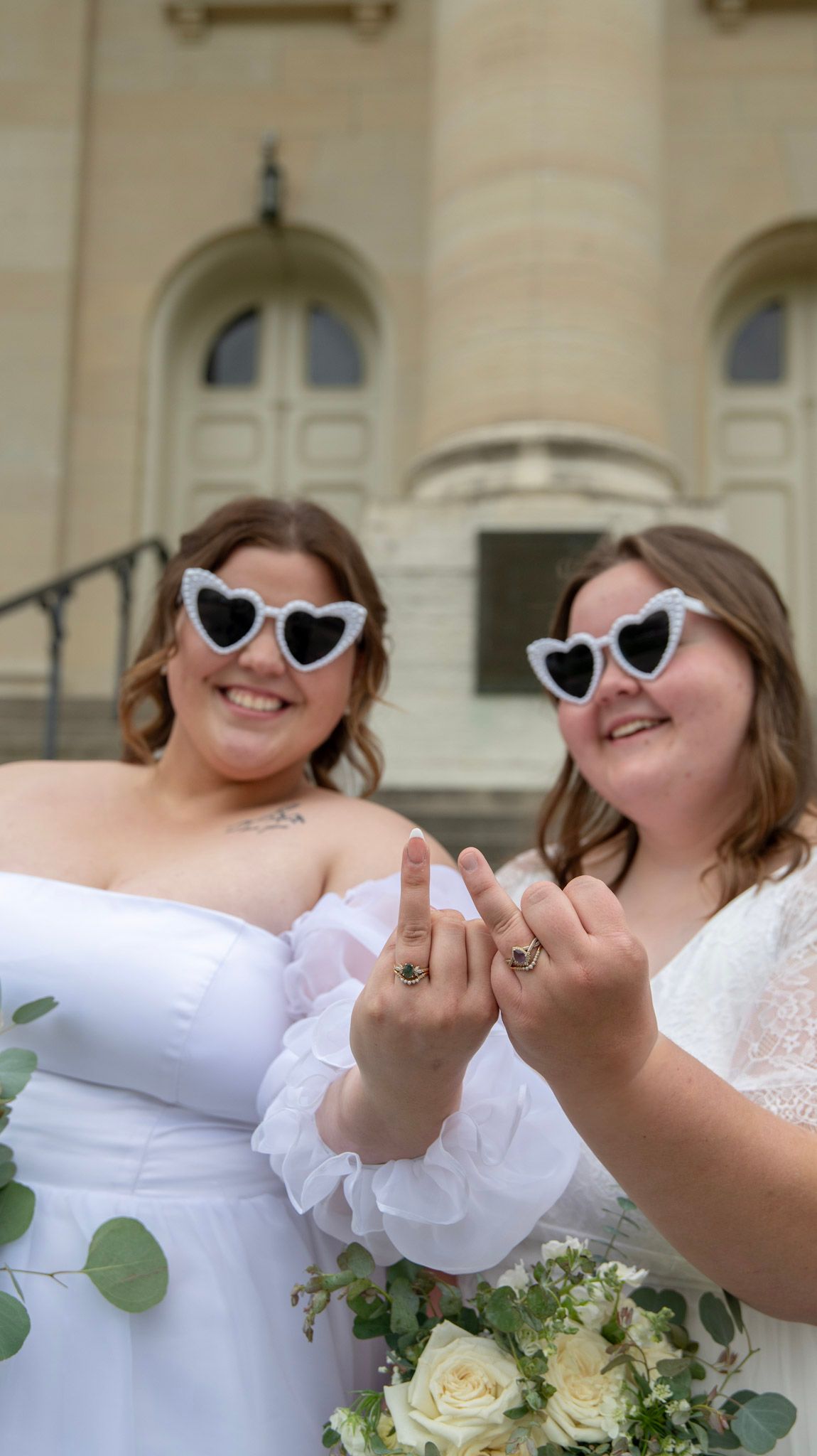 Newlyweds wearing heart-shaped sunglasses showing off rings with white bouquets.