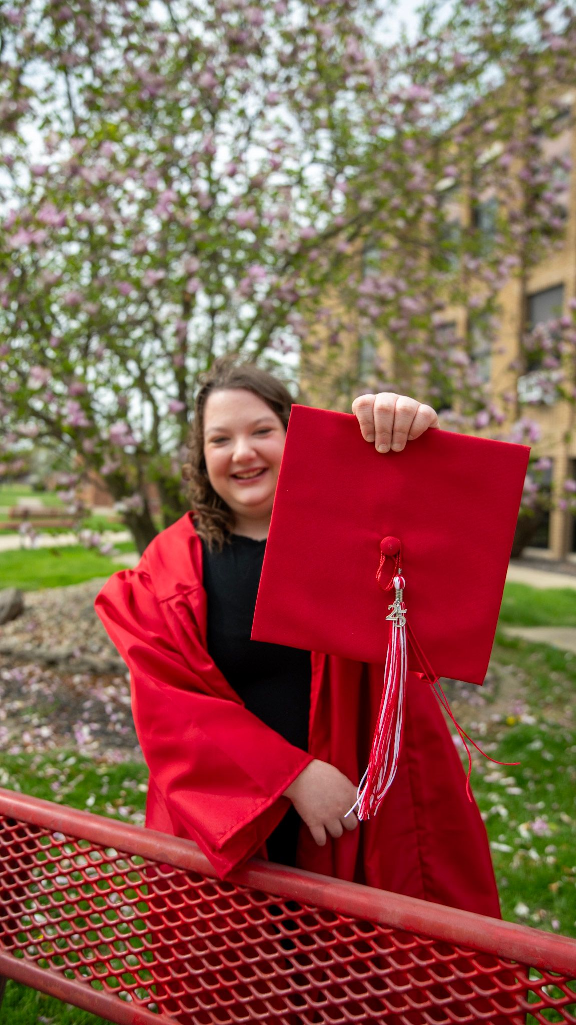 Graduate displays red diploma holder while leaning on railing with flowering trees in background.