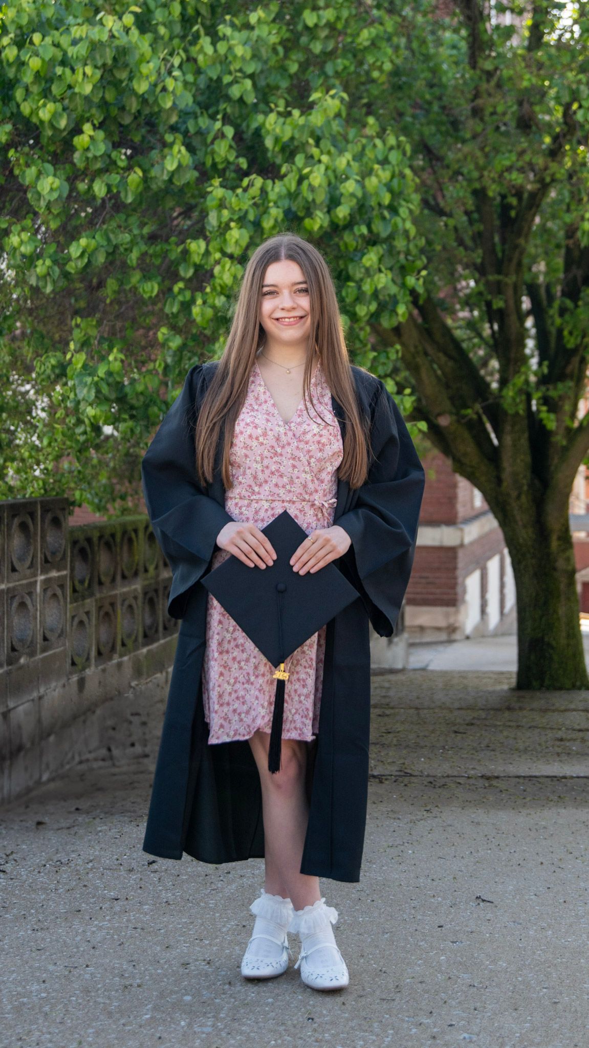 Graduate in floral dress and black gown poses playfully on stone pathway.