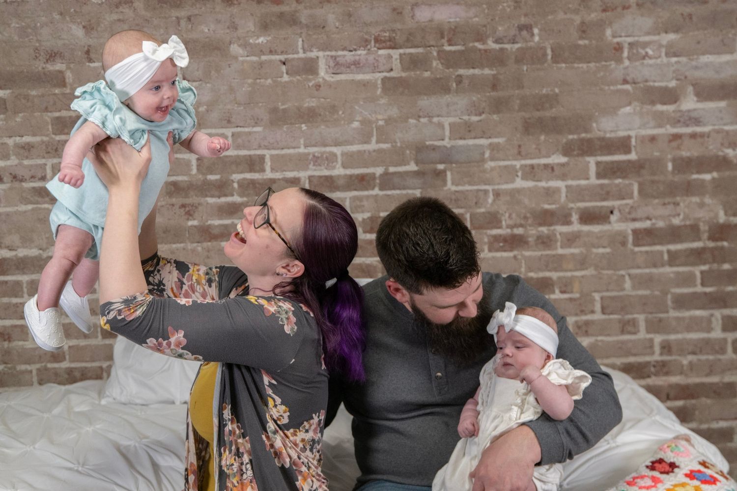 Parents share a playful moment with an infant on a couch against exposed brick.