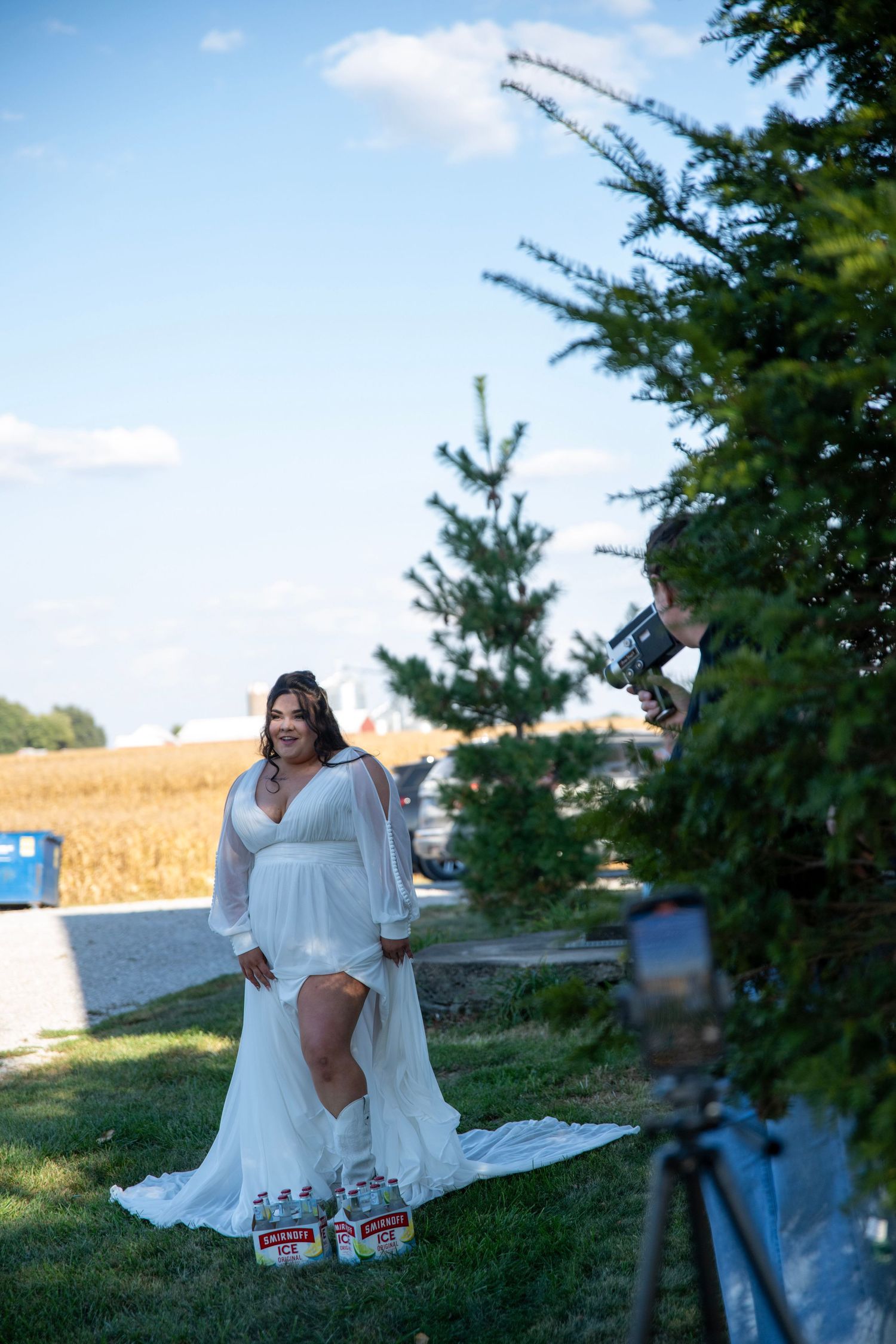 A person in a white dress poses in a grassy area with cornfields in the background.