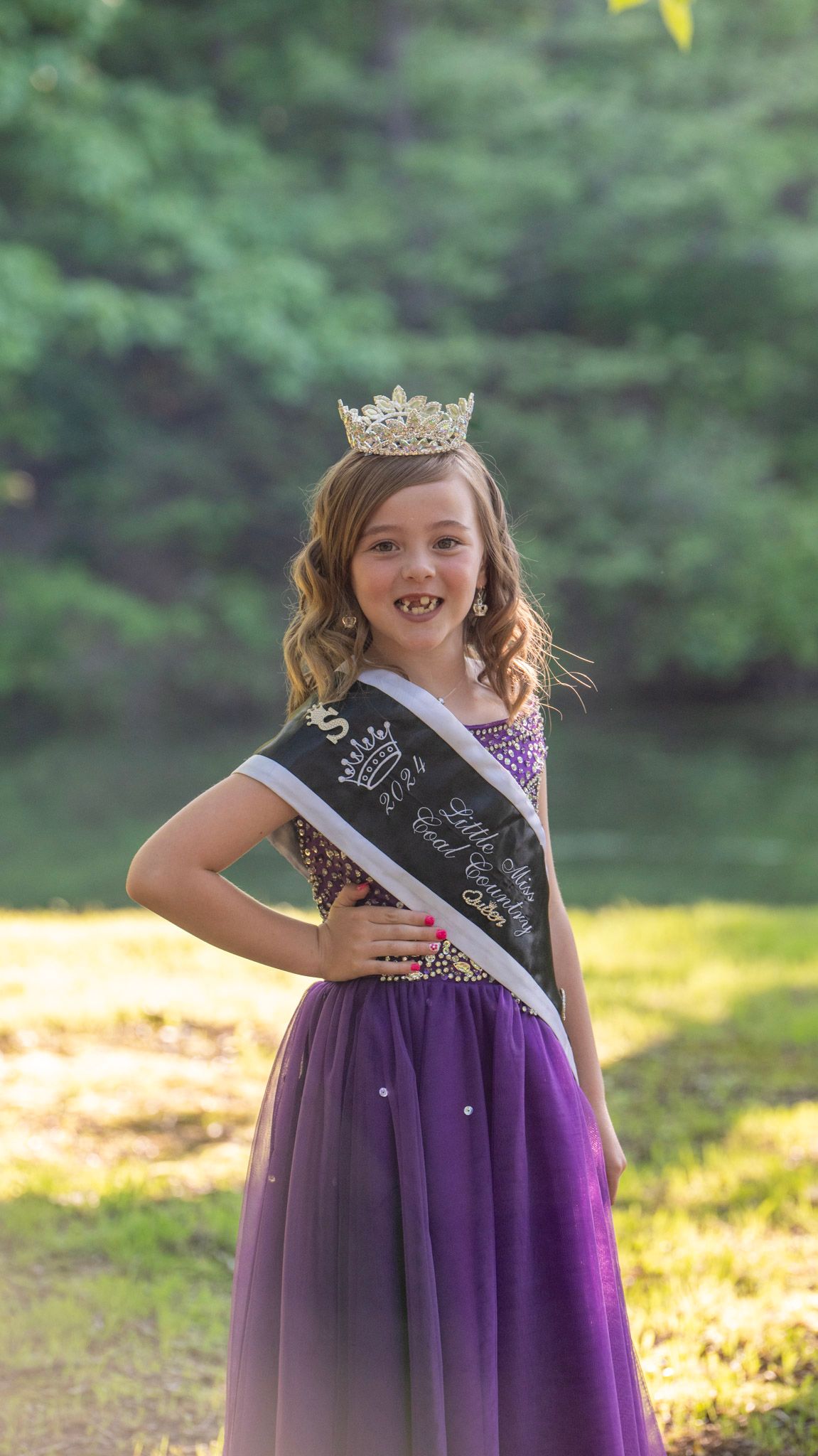 A contestant in ombre purple gown poses with crown and sash in a wooded setting.