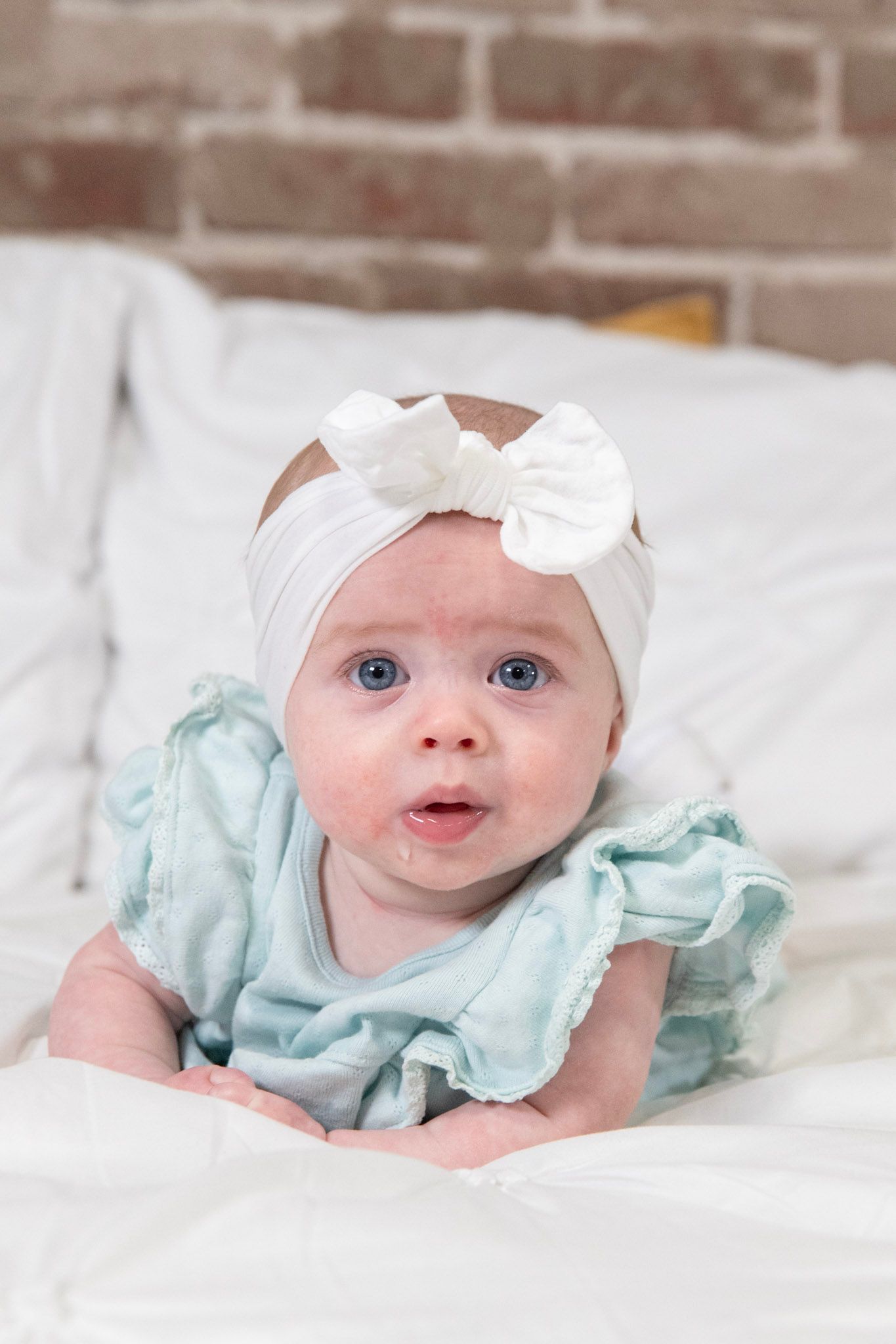 Baby in mint green ruffled dress and white bow headband lying on white bedding.