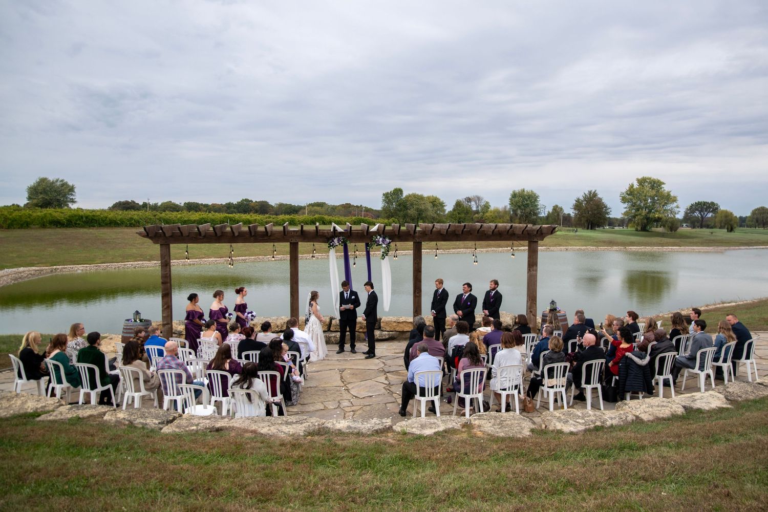 Aerial view of outdoor wedding ceremony by lake with wooden pergola and white chairs.