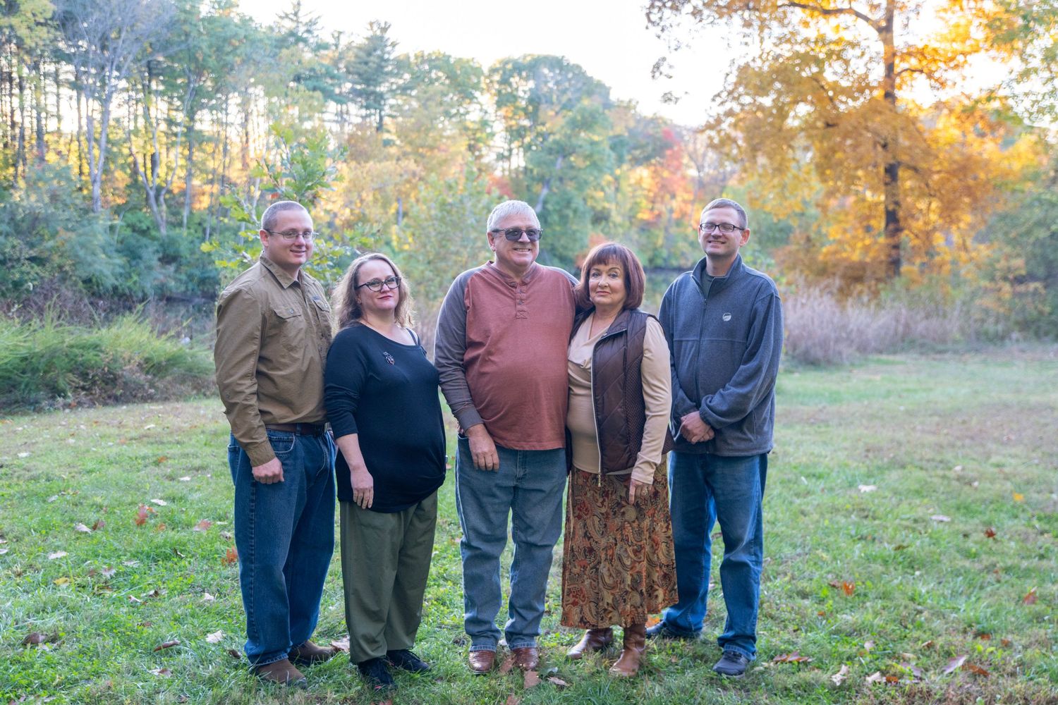 Family members stand together for group portrait in an autumn field with trees.