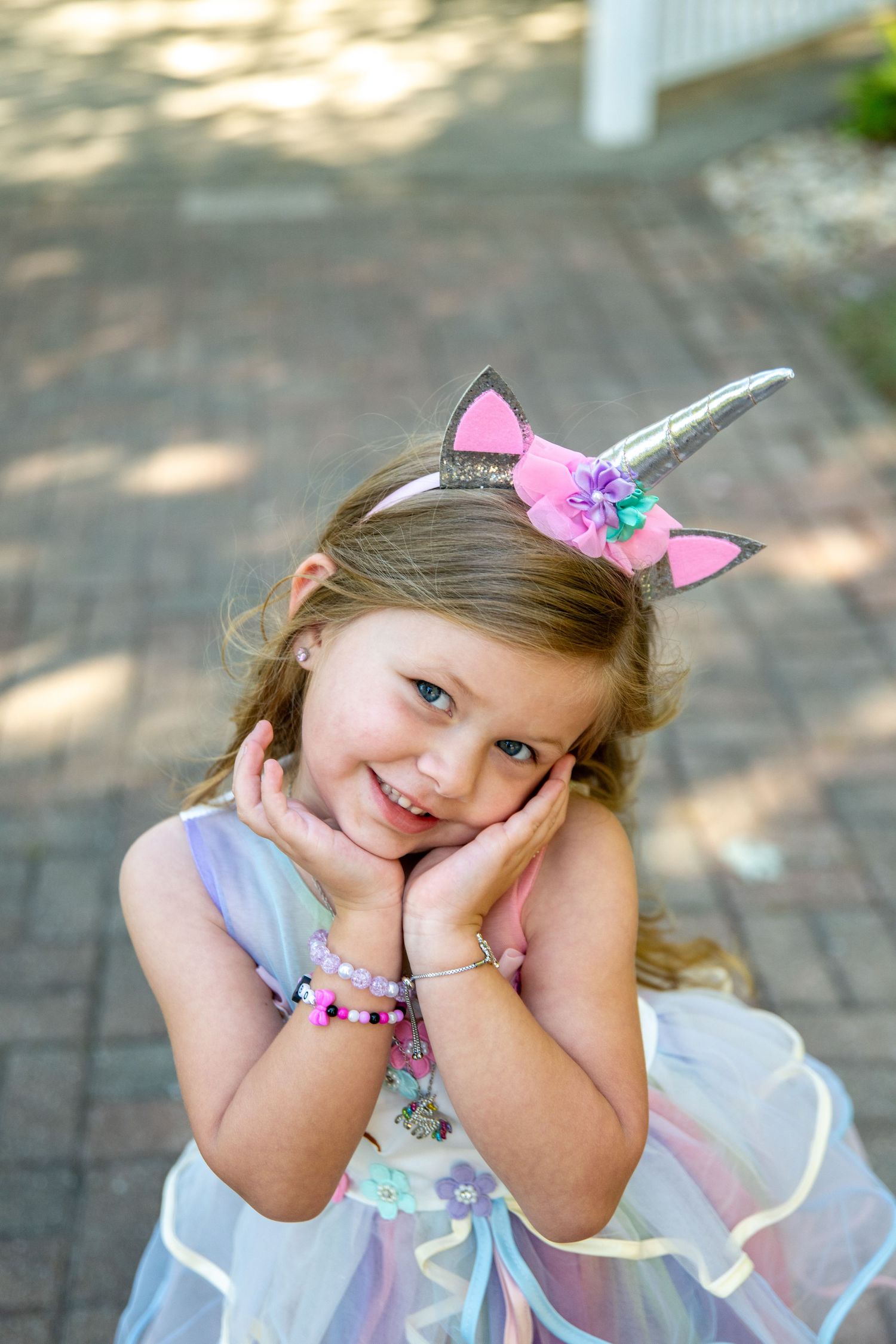 Child wearing unicorn accessories and pastel dress poses playfully on brick patio.