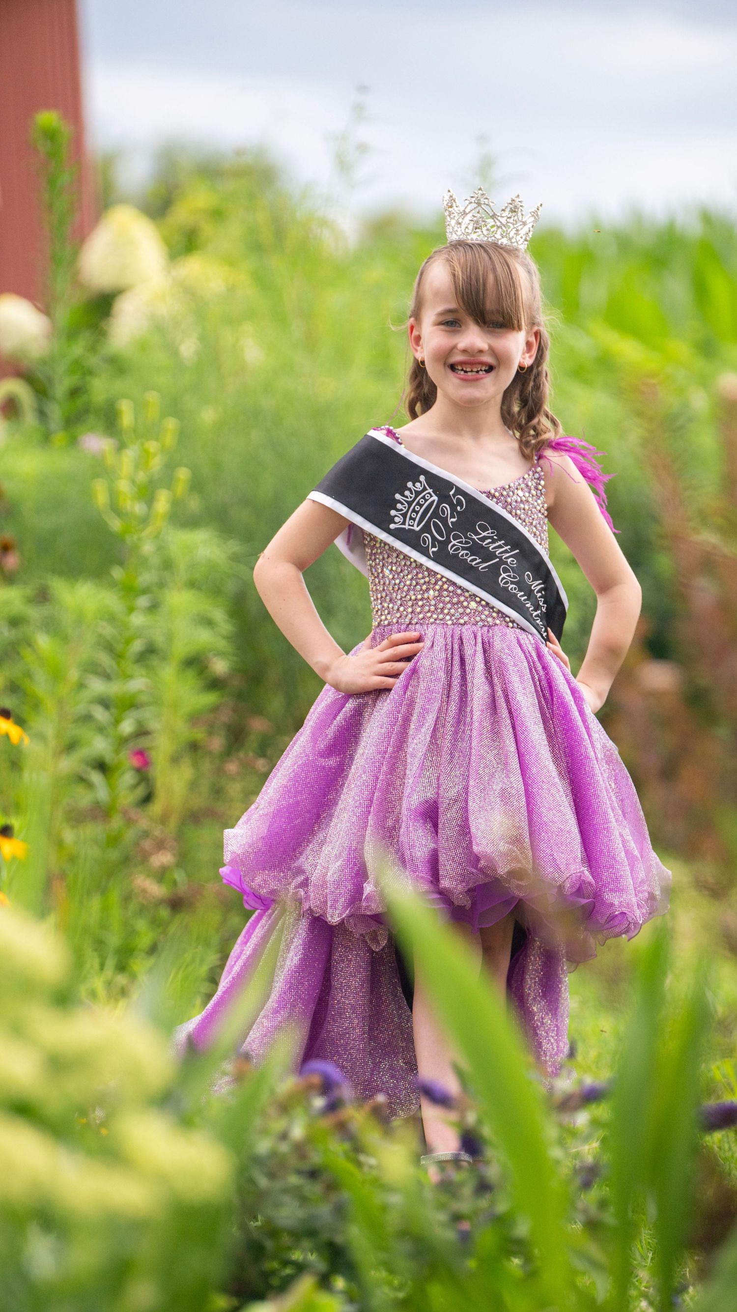 Young pageant winner in purple sparkly dress poses in garden setting wearing crown and sash.