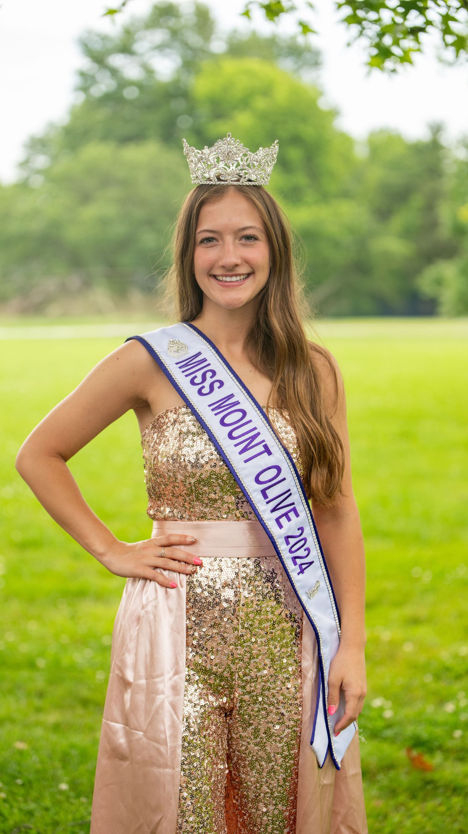 Pageant contestant wearing a gold sequined dress and crown posing in a grassy outdoor setting.